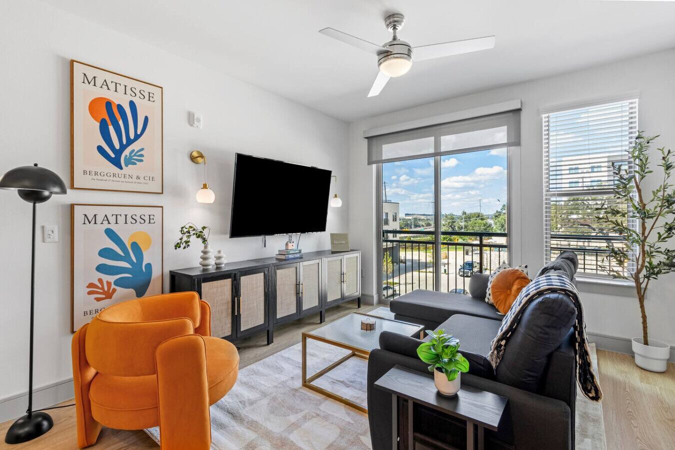 Een moderne woonkamer met een flatscreen televisie, oranje fauteuils, een zwarte bank, een plafondventilator en een balkon met uitzicht.