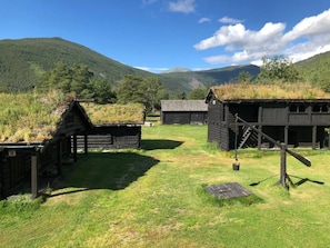 A rustic log cabin with a thatched roof, surrounded by a grassy field and mountains in the background.