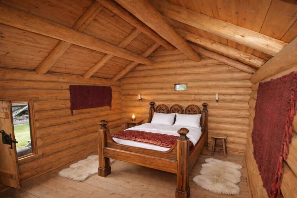 Ein Schlafzimmer in einer Blockhütte mit einem hölzernen Bett, einem kleinen Nachttisch, einem Fenster und einer Tür.