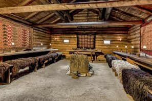 Interior de una cabaña de troncos con bancos, una mesa central y un tapiz decorativo en la pared.