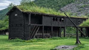 Une cabane en bois rustique, dotée d’un toit de chaume, d’un escalier menant à un étage supérieur et d’une petite terrasse.