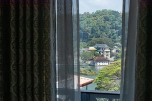 A view through a window with curtains, featuring a lake, buildings, and a forested hill. - Ceyloni Lake Residency - Kandy (Kandy)