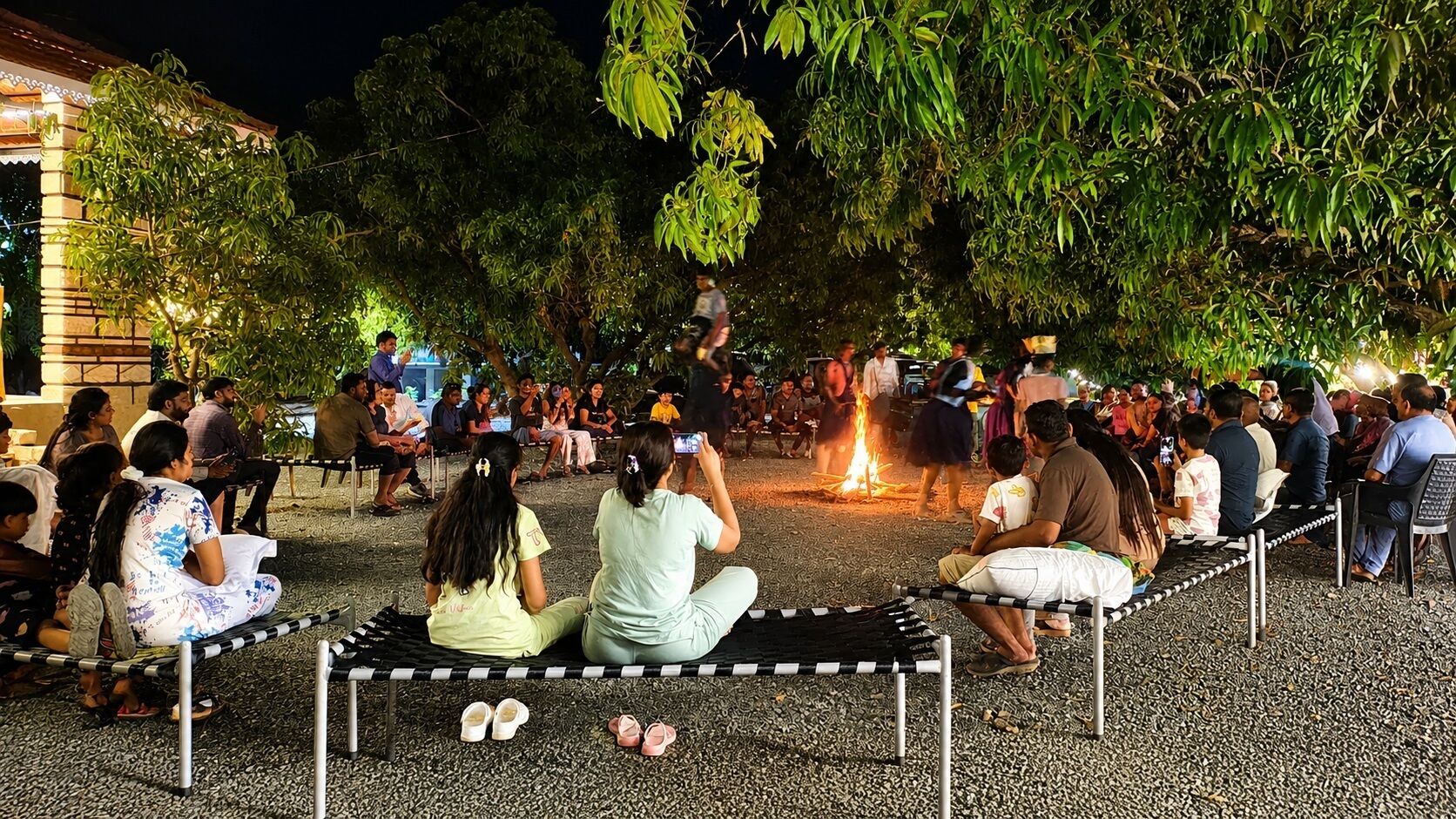 An outdoor gathering with people seated around a fire, trees, and a building in the background.