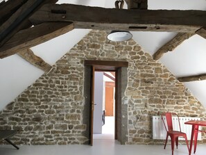 A stone-walled room with a sloped ceiling, a wooden beam structure, a table, and a chair.