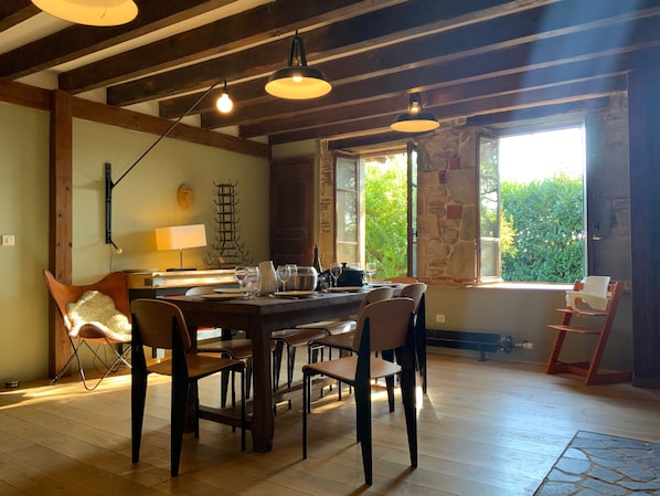 A dining area with a wooden table, chairs, pendant lights, a stone wall, and a window with a view of greenery.