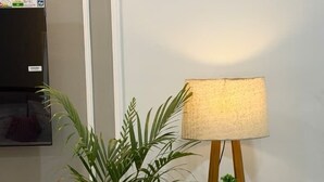 A potted plant next to a wooden floor lamp with a white shade, a small shelf with two plants, and a TV mounted on the wall. - Supreme Stays (Indore)