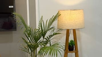 A potted plant next to a wooden floor lamp with a white shade, a small shelf with two plants, and a TV mounted on the wall.