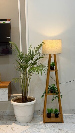 A potted plant next to a wooden floor lamp with a white shade, a small shelf with two plants, and a TV mounted on the wall. - Supreme Stays (Indore)