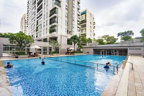 En swimmingpool omgiven av en modern byggnad med balkonger, ett takförsett sittområde och en klar himmel.