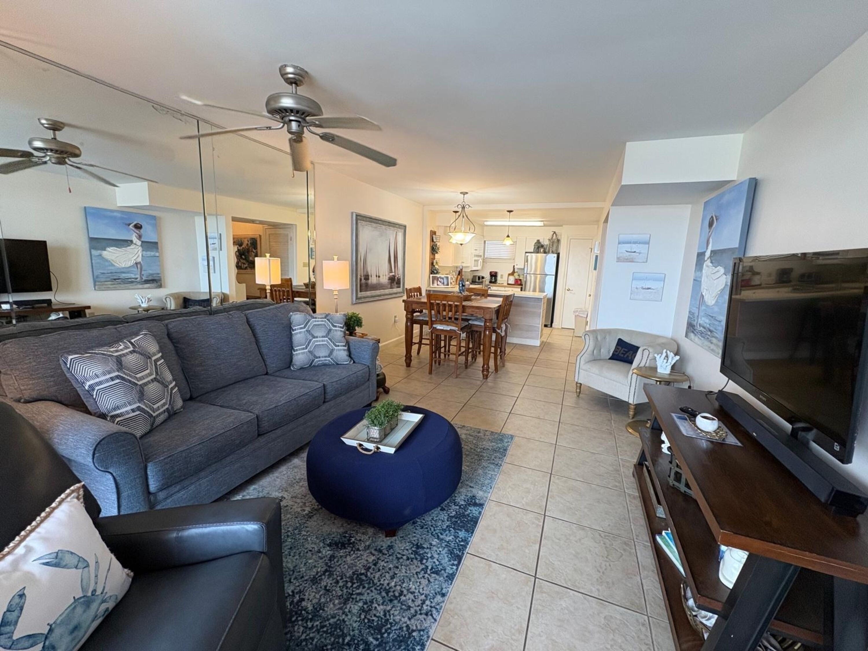 A modern living room with a grey sofa, a blue ottoman, a TV, and a dining area with wooden furniture.