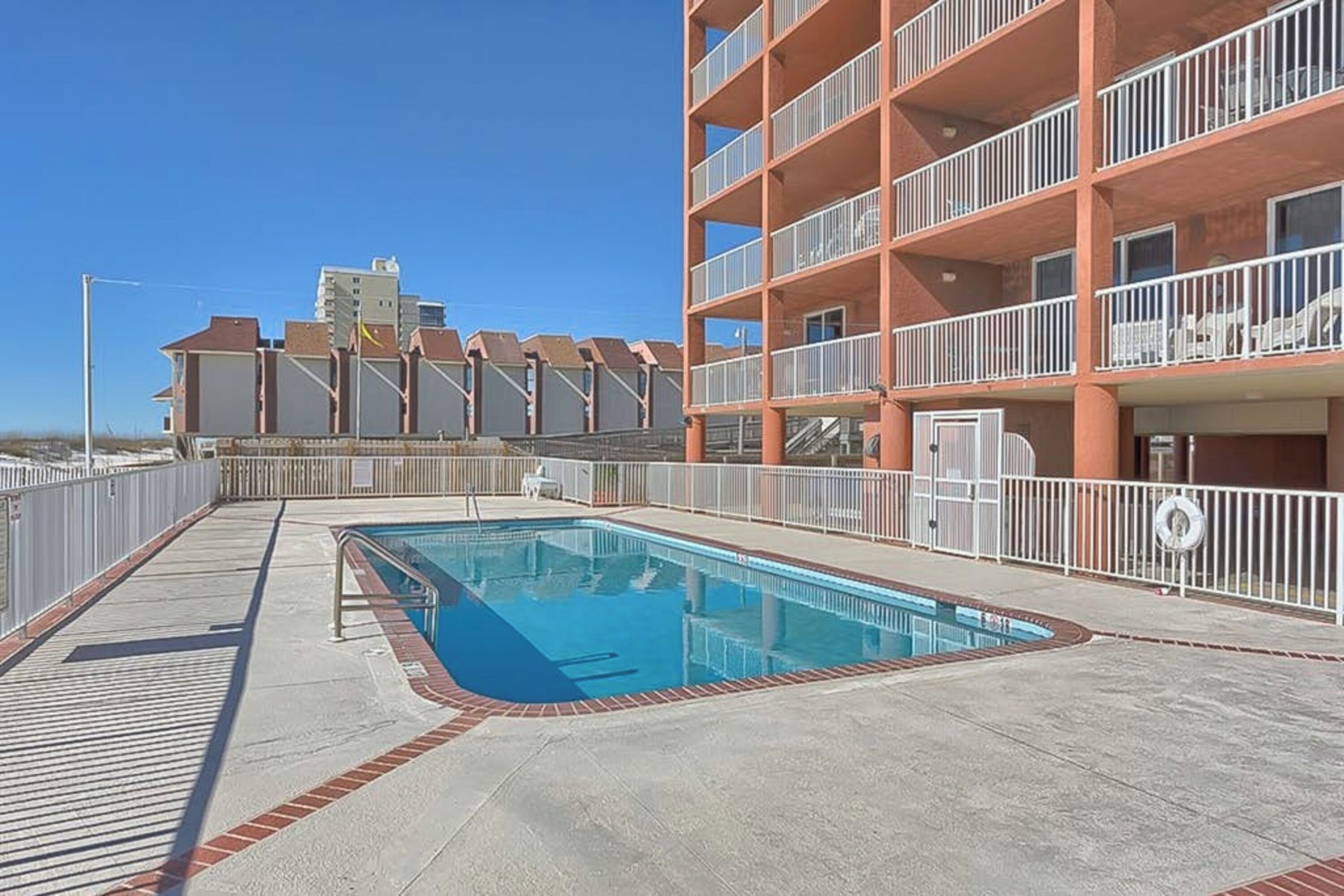 A rooftop pool area with a clear blue pool, a white railing, and a building with balconies in the background.