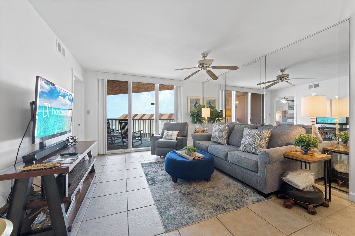 A modern living room with a sofa, ottoman, TV, ceiling fans, and a balcony with a view.