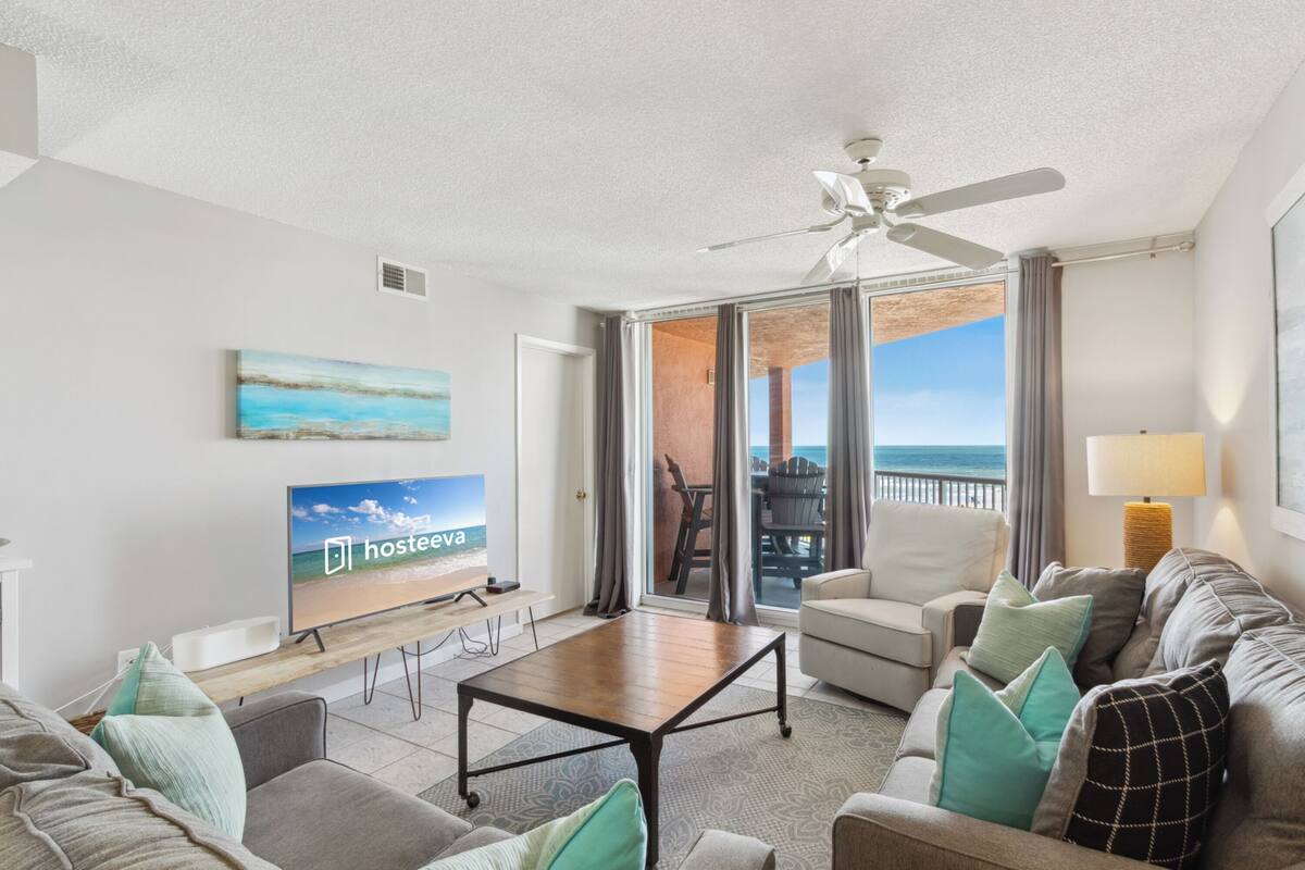 A modern living room with a flat-screen TV, a ceiling fan, and a view of the ocean through large sliding doors.