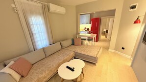 A living room with a sofa, a small round table, and a kitchen area in the background. - Jasmine Suıts (ula)
