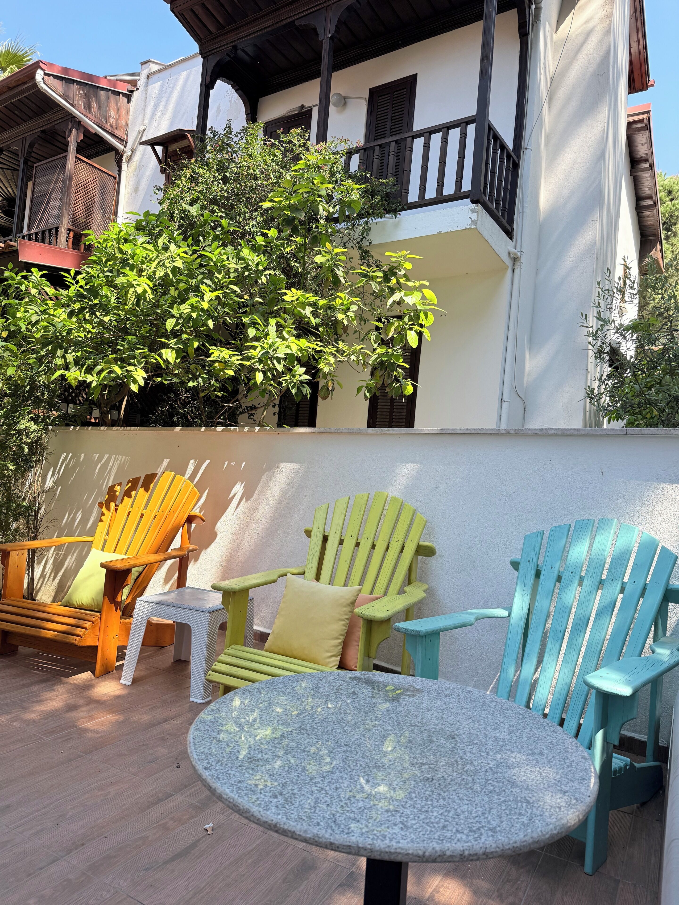 A patio area with wooden chairs and a table, surrounded by a white wall and greenery.