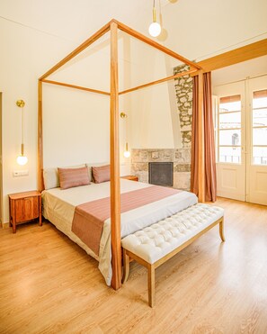 Uma cama com dossel, uma lareira de pedra, soalho de madeira e um banco.