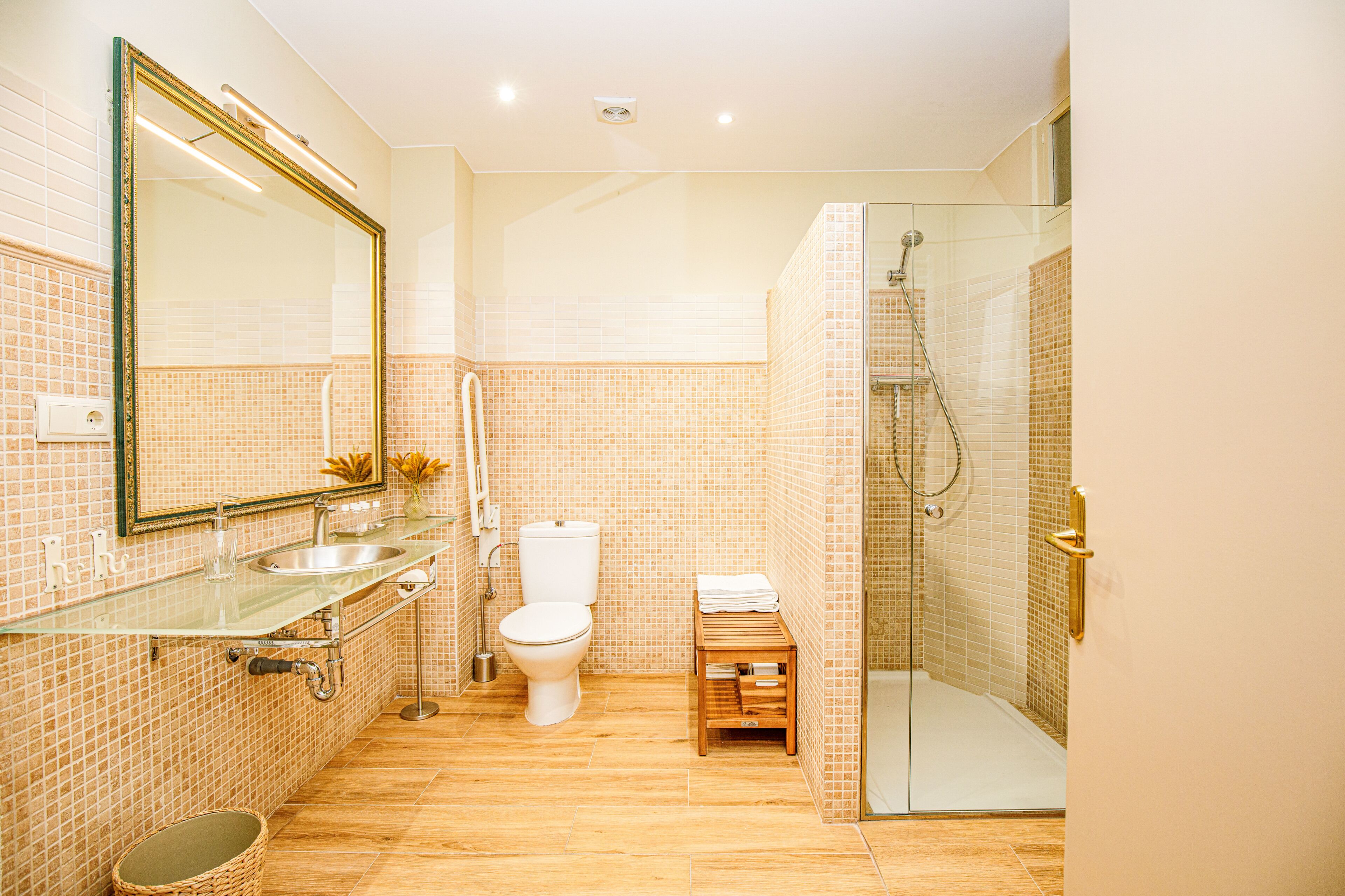 A bathroom with a glass shower enclosure, a toilet, a sink with a glass countertop, and a mirror with a gold frame.