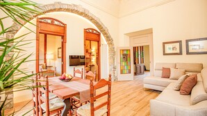 A dining area with a wooden table and chairs, a stone archway, a living room with a sofa, and a television. - LITTLE PALACE SWIMING POOL Unique in the center (Besalu)