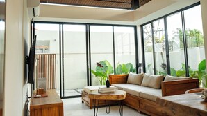 Ein modernes Wohnzimmer mit einer Couch, einem Couchtisch und einem Fernseher. Große Fenster und ein Blick ins Grüne sind zu sehen.
