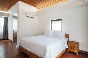 Un dormitorio con cama de madera, ropa de cama blanca, una mesita de noche, una ventana y un equipo de aire acondicionado.