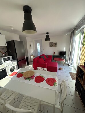A modern kitchen with a washing machine, a dining area with a table set for two, a red sofa, and a television in the living area. - La Providence (La Tremblade)