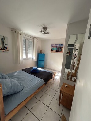 A bedroom with a bed, a blue dresser, a window with blinds, a ceiling fan, and a mirror. - La Providence (La Tremblade)