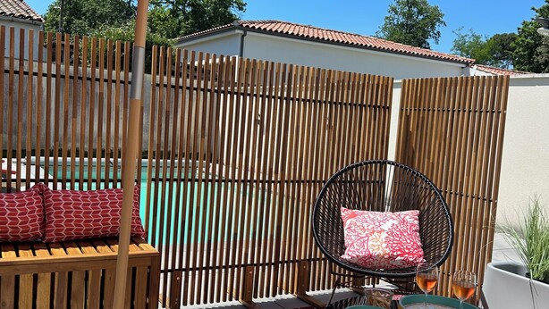Eine Terrasse mit Holzlattenzaun, einem roten Sonnenschirm, einem Korbstuhl, einem kleinen Tisch mit Gläsern und einer Bank mit gemusterten Kissen.