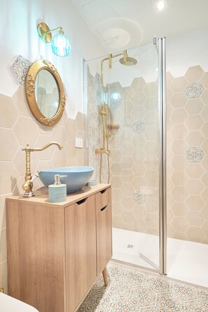 A bathroom with a wooden vanity, a blue vessel sink, a gold-framed mirror, a shower with a glass door, and hexagonal patterned walls. - La Dimora Blu (Albenga)
