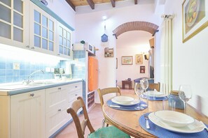 Un comedor con una mesa de madera dispuesta para cuatro, una cocina con armarios blancos y azulejos azules, y un arco de ladrillo que conduce a otra habitación.