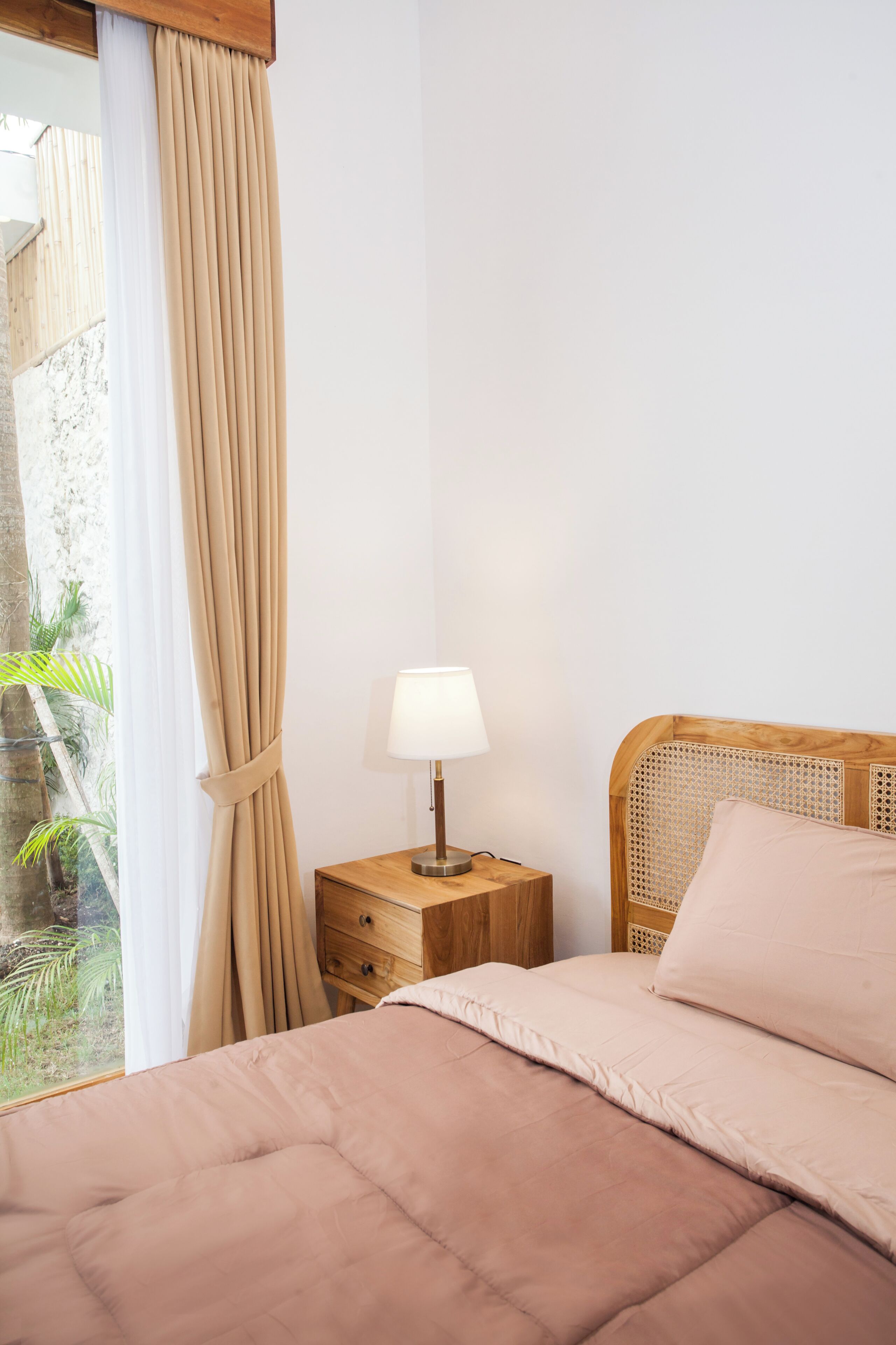 Een slaapkamer met een bed, een nachtkastje met een lamp, een raam met gordijnen en uitzicht op groen buiten.
