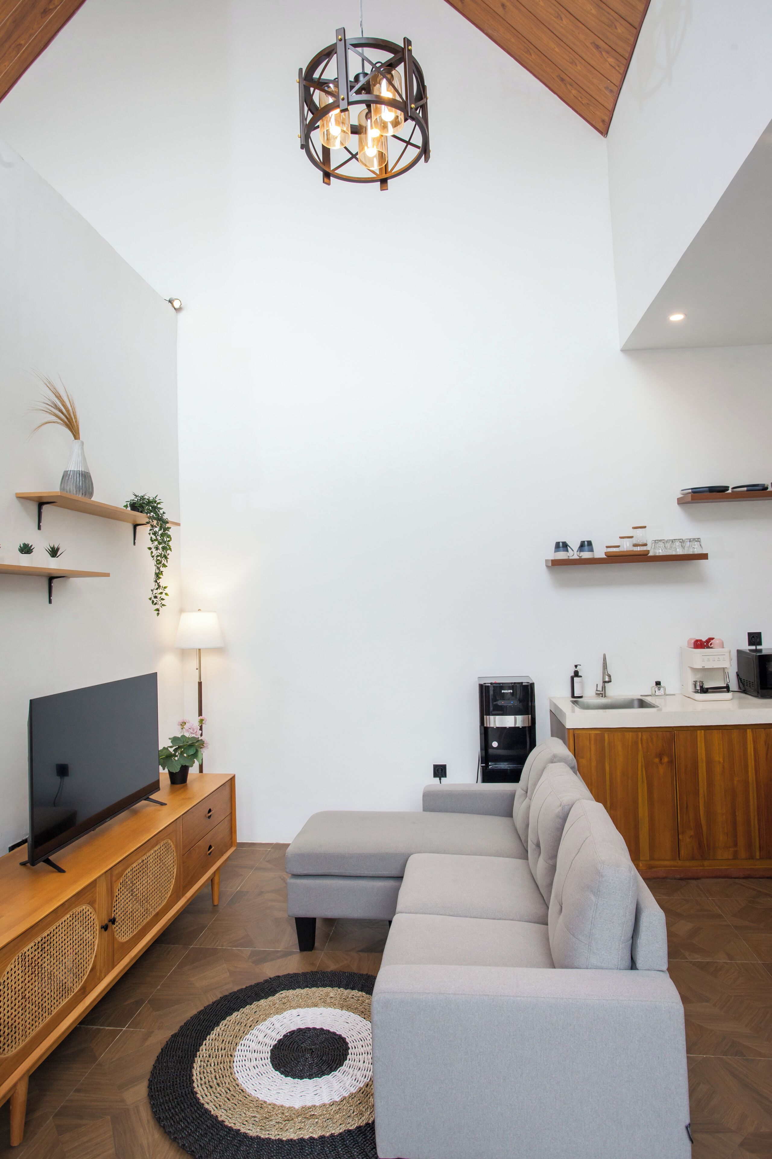 Una sala de estar moderna con un sofá gris, unidad de entretenimiento de madera y una zona de cocina con encimera blanca y armarios de madera.