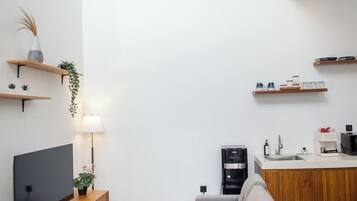 Una sala de estar moderna con un sofá gris, unidad de entretenimiento de madera y una zona de cocina con encimera blanca y armarios de madera.
