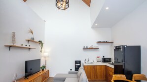 Un espacio habitable moderno con un televisor de pantalla plana, un sofá seccional gris, una mesa de comedor de madera, una cocina con un refrigerador negro y un mueble de madera empotrado.