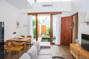 A modern living room with a sofa, dining table, chairs, a TV, and a sliding glass door leading to a garden. - Private Pool 2-BR Villa in Seminyak - C1 (Denpasar)