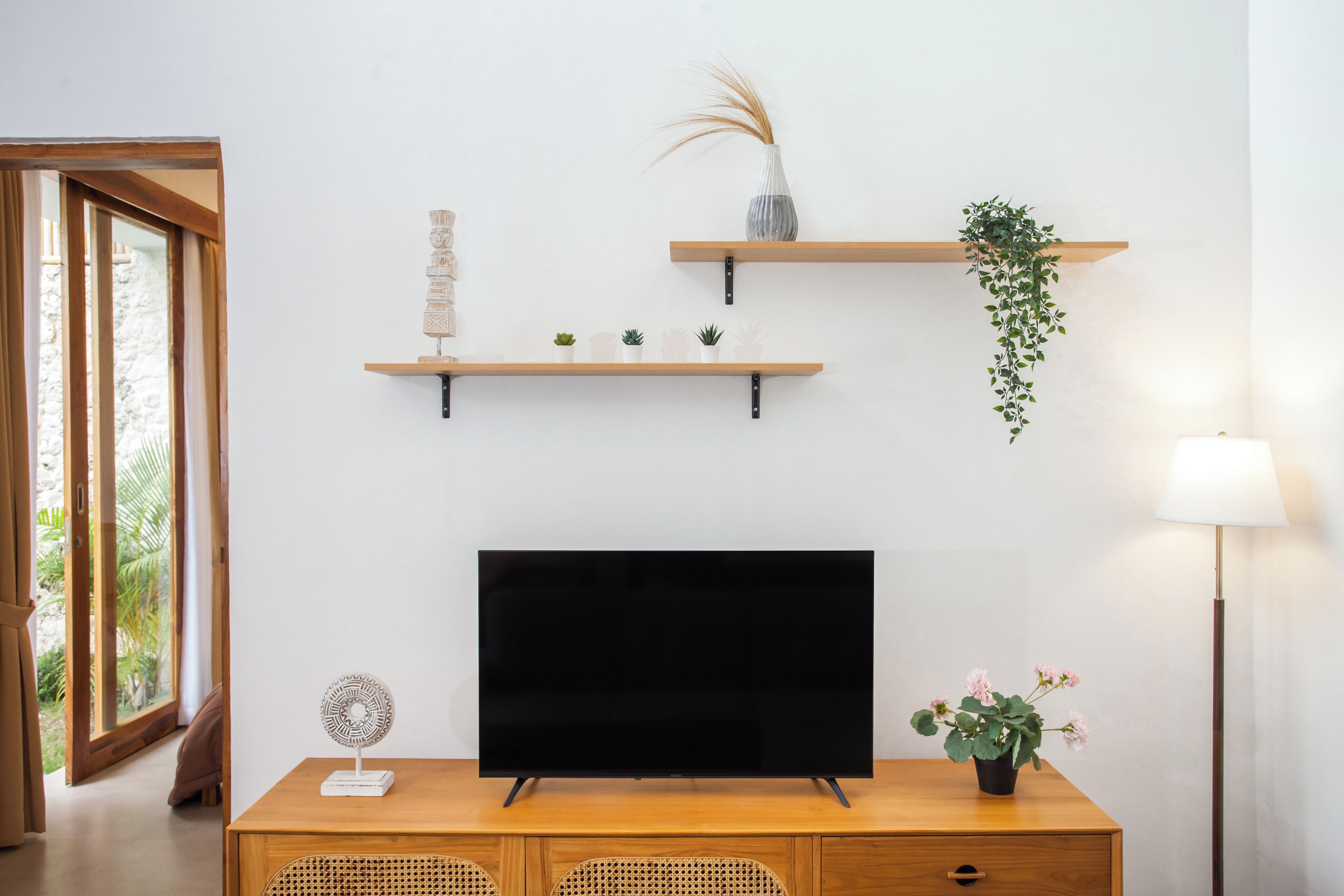Una sala de estar moderna con una televisión de pantalla plana, estanterías de madera con objetos decorativos, una planta en maceta y una lámpara de pie.