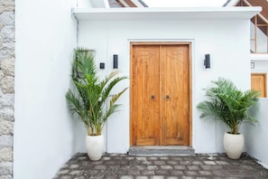 A white building with a wooden double door, two potted plants, and a stone wall. - Private Pool 2-BR Villa in Seminyak - C1 (Denpasar)