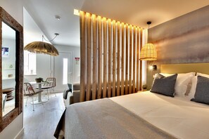 Quarto de hotel moderno com uma cama grande, área de refeições com cadeiras, divisória de madeira e luminária pendente.