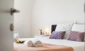 Ein ordentlich bezogenes Bett mit aufgerollten Handtüchern, ein Nachttisch mit einer Pflanze und eine Wandleuchte.