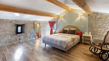 Une chambre avec un mur en pierre, des poutres en bois, un lit, un fauteuil à bascule et une petite fenêtre.