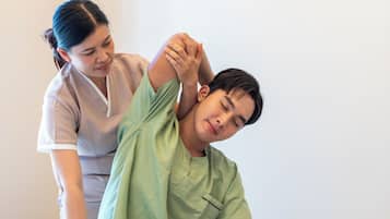 A person receiving a physical therapy session on a bed in a room with a plain wall and a white pillow.