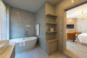 A modern bathroom with a freestanding bathtub, wooden shelves, a vanity, and a television in the adjacent bedroom. - Kaysorn Siri (Sirindhorn)