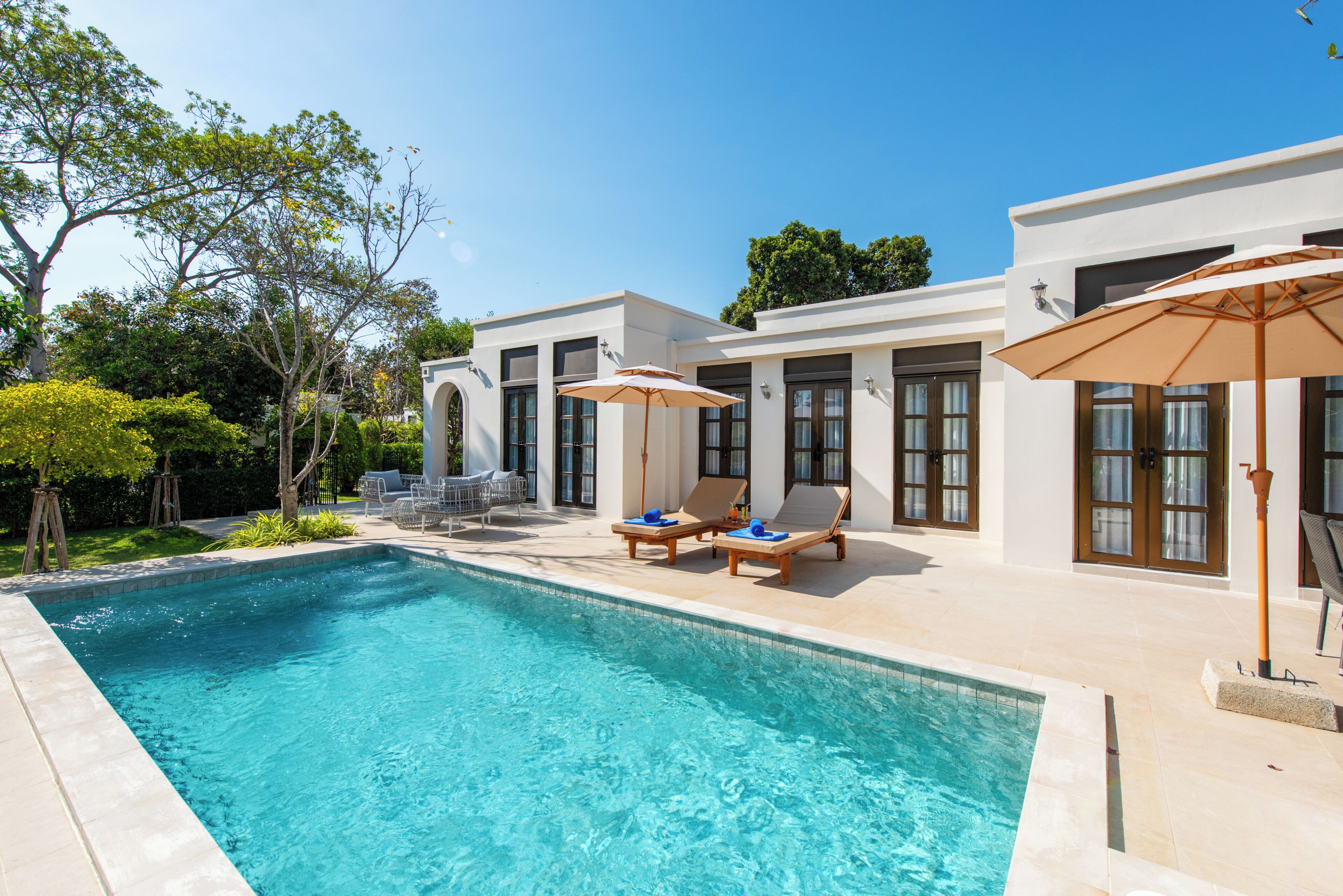 Una piscina con sillones de descanso y sombrillas, rodeada por un edificio blanco con amplias ventanas y puertas.