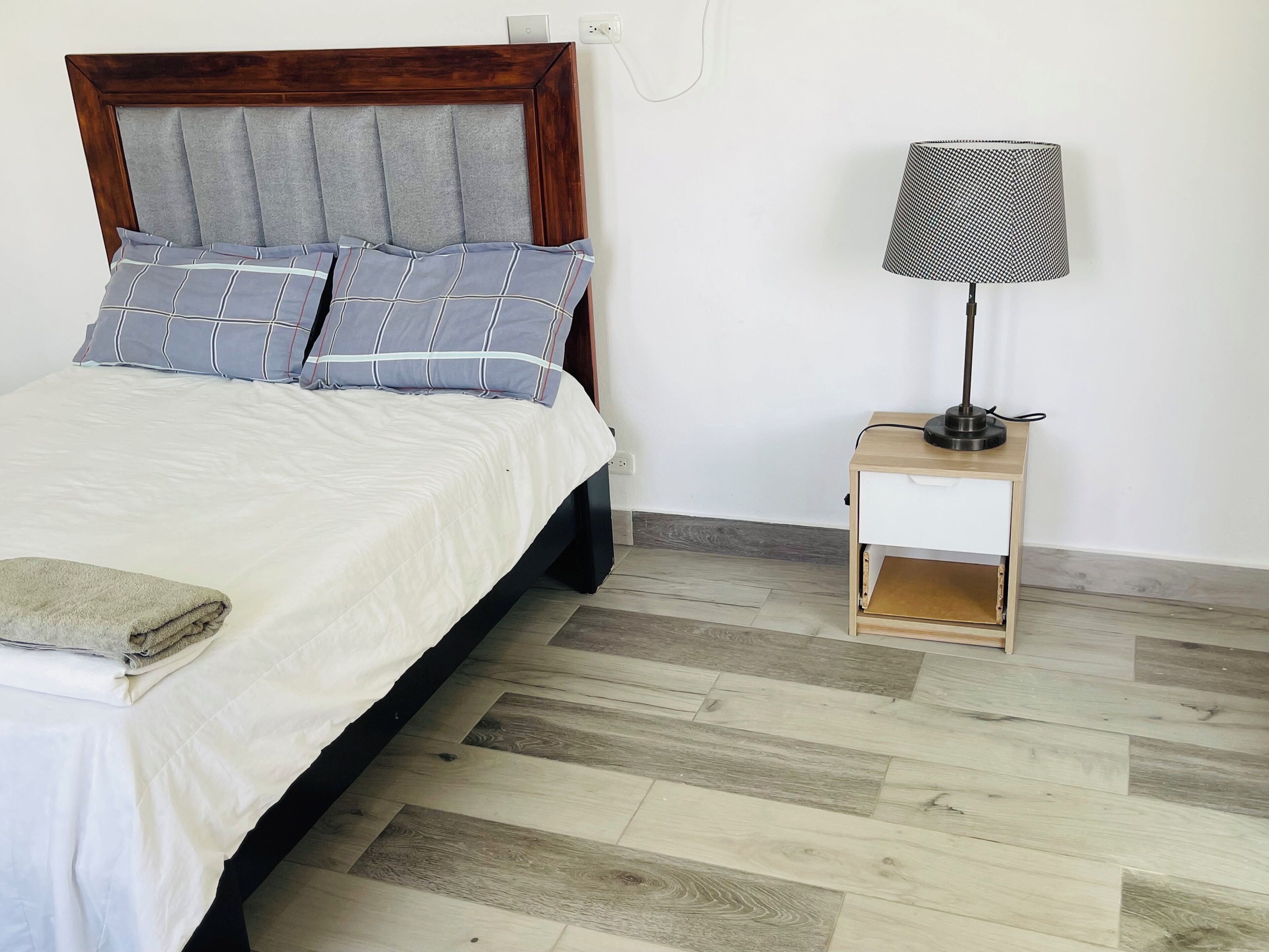 A bed with a wooden headboard, grey and blue striped pillows, a white bedspread, a folded towel, a nightstand with a lamp, and a wooden floor.
