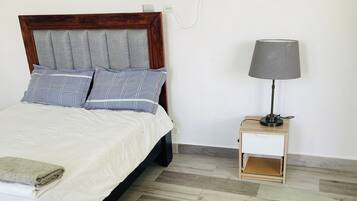 A bed with a wooden headboard, grey and blue striped pillows, a white bedspread, a folded towel, a nightstand with a lamp, and a wooden floor.