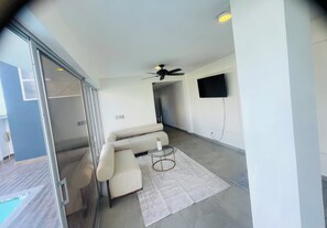 A modern living room with a sofa, a glass coffee table, a ceiling fan, and a flat-screen TV mounted on the wall.