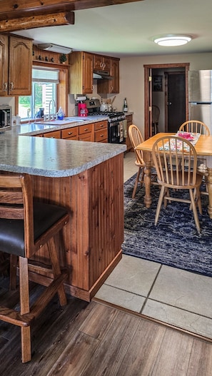 A kitchen with wooden cabinets, a granite countertop, a dining table with chairs, and a rug. - Bell Inn Cottage - Sanford Lake (Sanford)