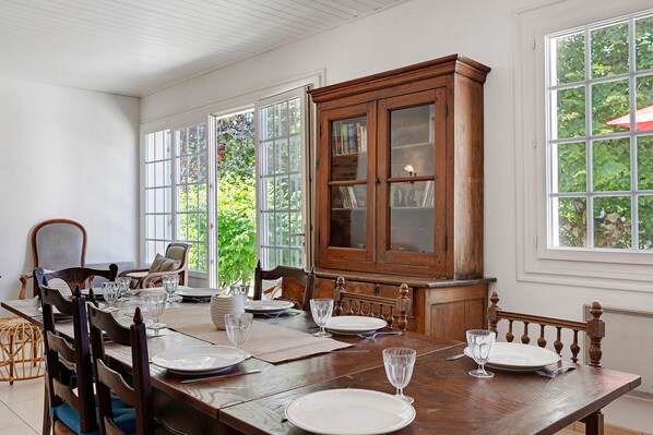 Une salle à manger avec une table en bois dressée pour six personnes, entourée de chaises, d’un buffet en bois et de grandes fenêtres.