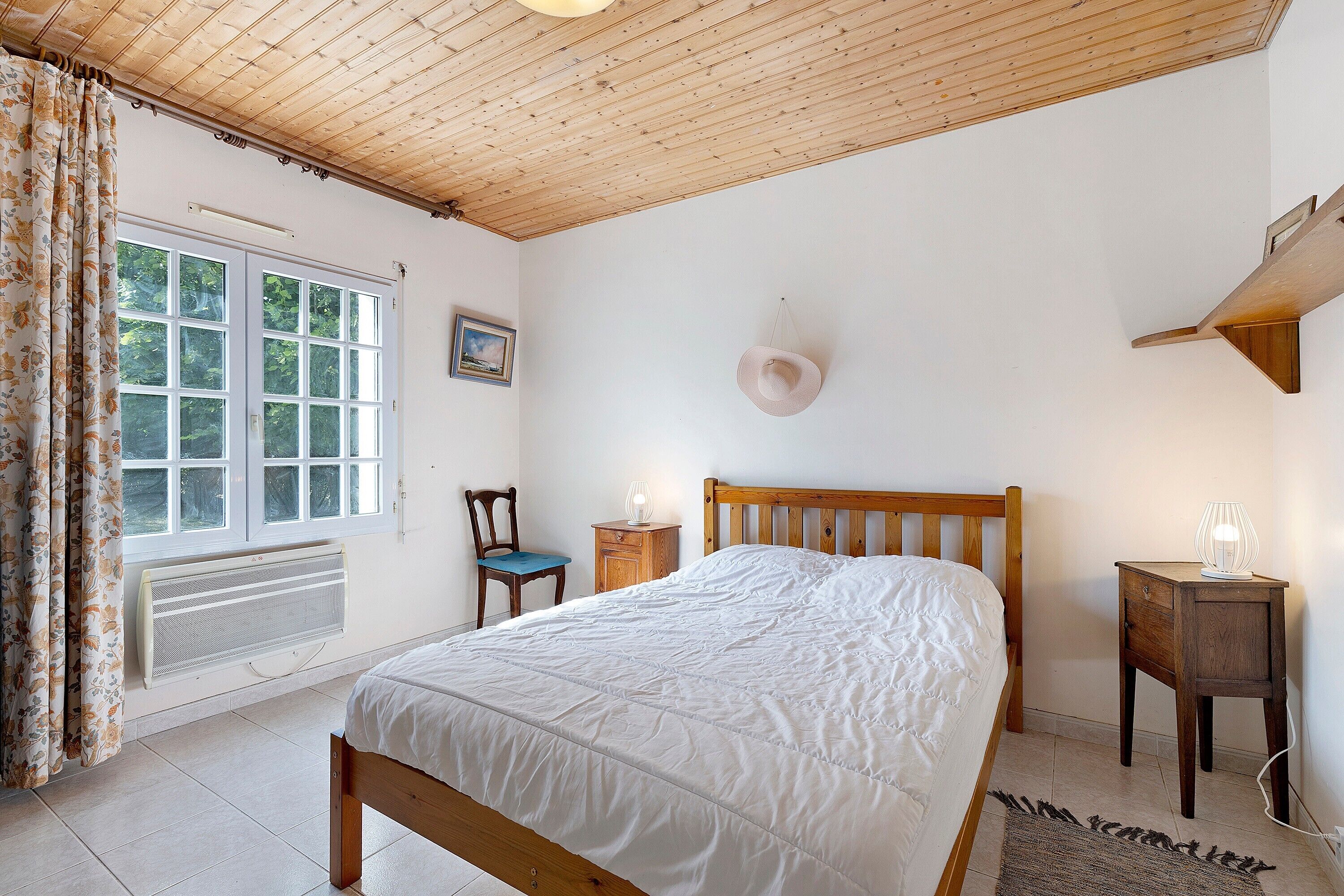 Ein Schlafzimmer mit Holzboden, einem Bett, einem Sessel, einem Nachttisch, einem Heizkörper und einem Fenster mit Vorhängen.