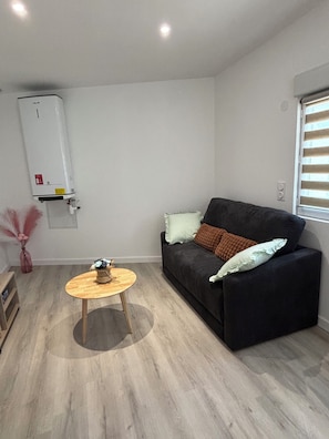 Een woonkamer met een zwarte bank, een ronde houten tafel, een witte radiator en een raam met zonwering.