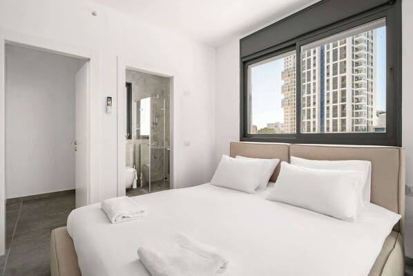 A modern hotel room with a large bed, a window with a city view, and a separate bathroom. - Modern 4BR Balcony Mamad Renovated Bldg (Bat Yam)